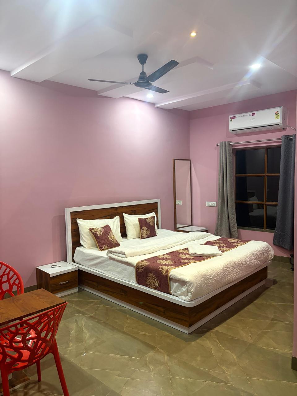 Guest room with soft pink walls, a double bed with maroon-and-gold runner, a split air-conditioner and a window with grey curtains